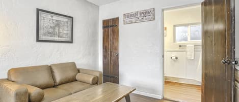 Una habitación con piso de madera, un sofá de cuero, una mesita de centro de madera, un mapa enmarcado y una puerta que da a un baño.