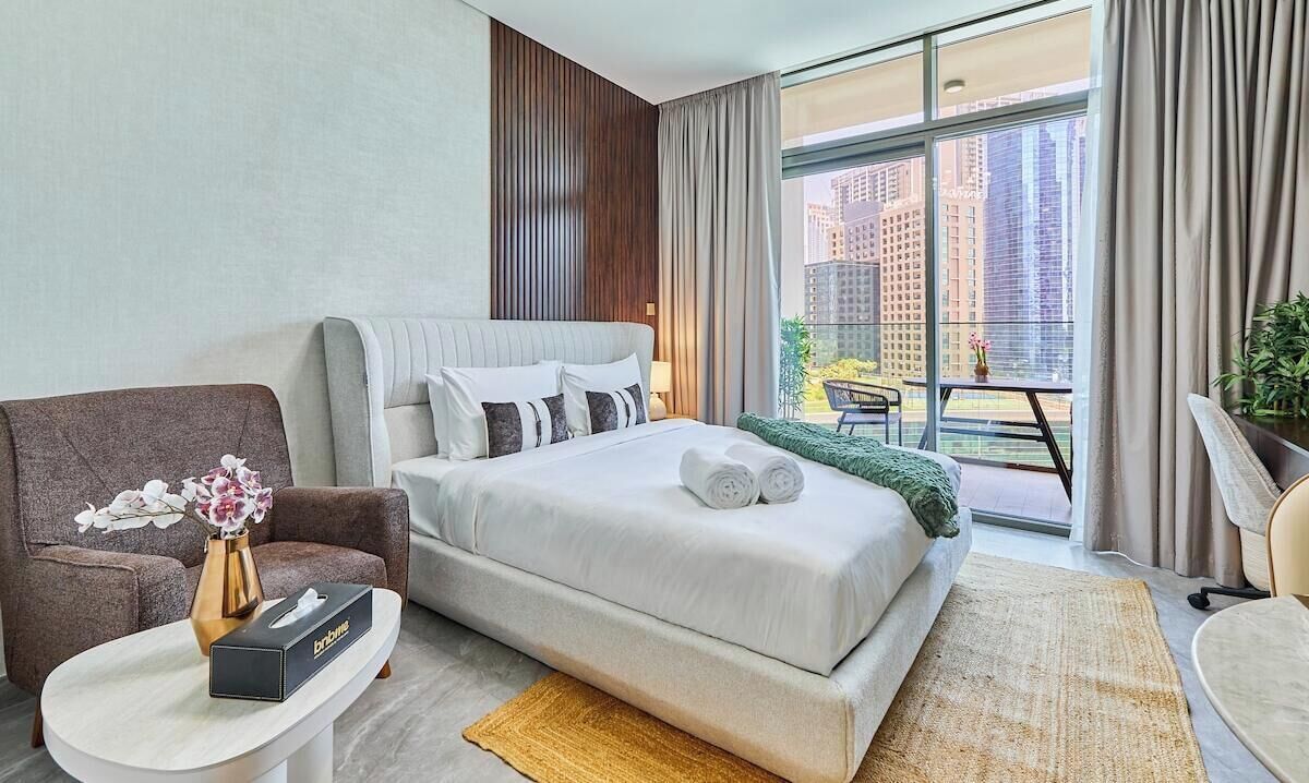 A hotel room with a bed, a chair, a small table with a vase, and a view of the city through large windows.