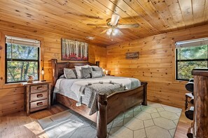 Une chambre à coucher dans un chalet en bois, avec un lit, des tables de chevet, un ventilateur de plafond et un tableau représentant une forêt.