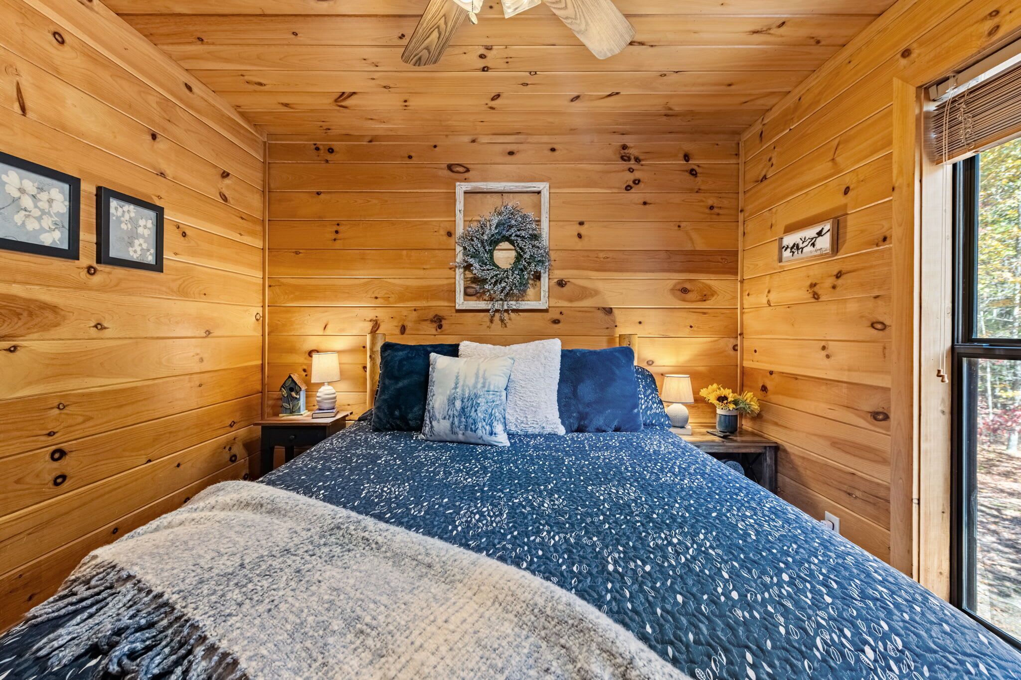 A wooden cabin bedroom with a bed, two nightstands, a wall-mounted lamp, a wreath, and a window with a view of trees.