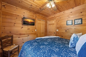 Ein Zimmer in einer Holzhütte mit einem Bett, einem Sessel, einem Deckenventilator und einem gerahmten Bild an der Wand.