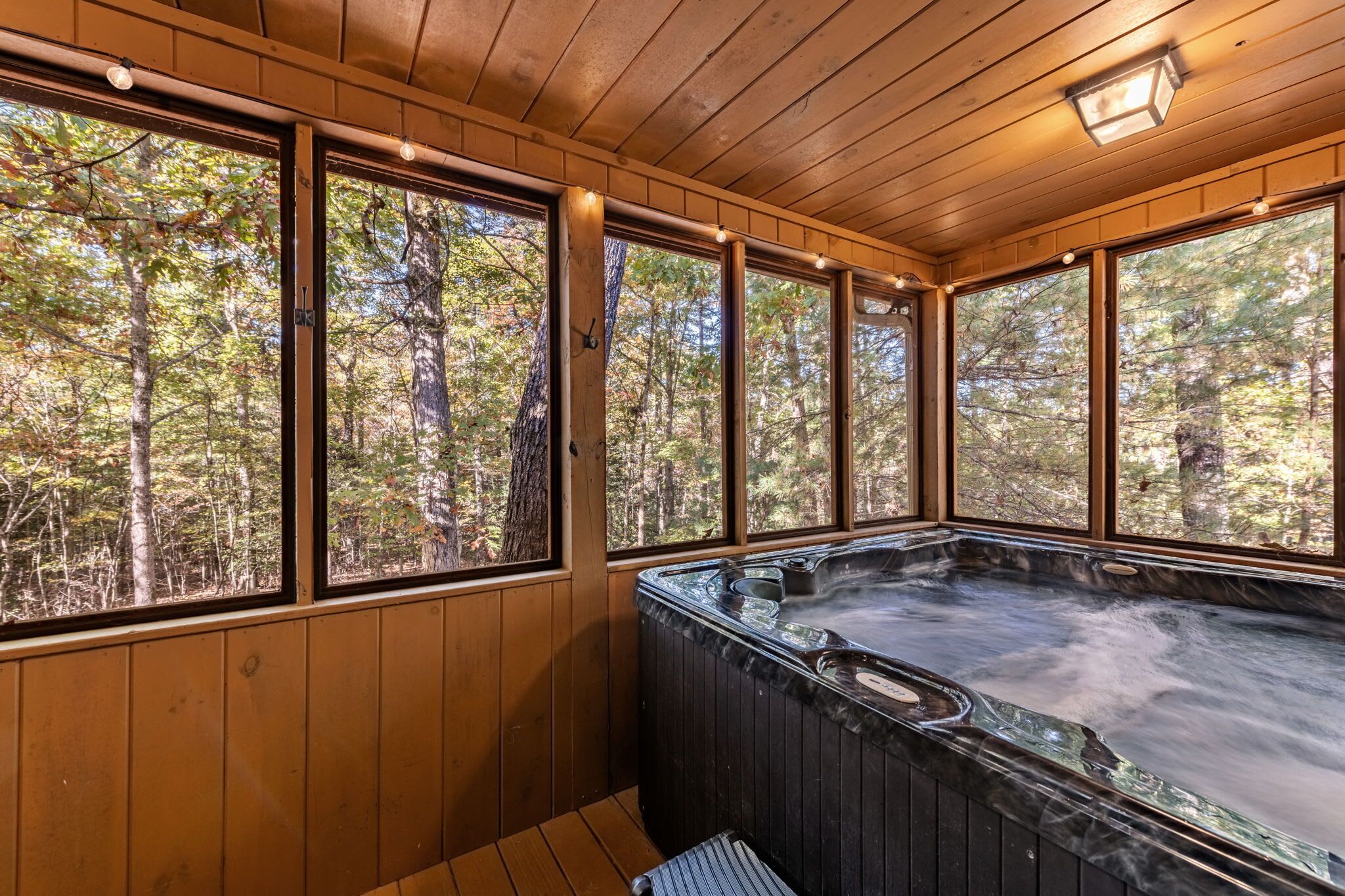 A wooden cabin with a hot tub and large windows overlooking a forest.