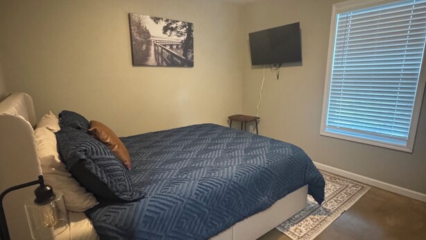 Ein Schlafzimmer mit einem Bett, einem an der Wand befestigten Fernseher, einem Fenster mit Jalousien und einer Deckenleuchte.