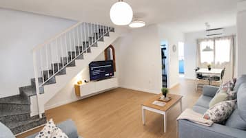 A modern living room with a staircase, a flat-screen TV, a dining area, and a sofa.