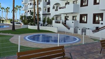 Un área de piscina con un banco de madera, rodeada por una valla, y un edificio con balcones al fondo.