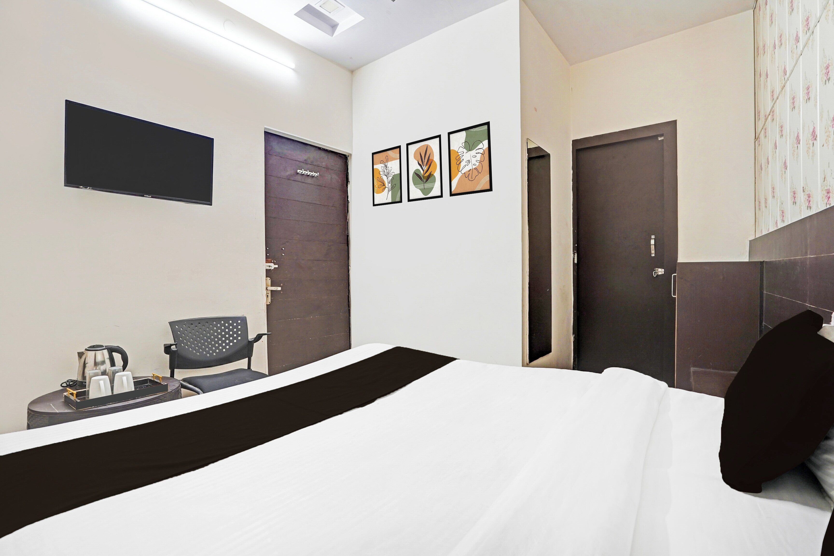 A hotel room with a bed, a chair, a small table with a kettle and cups, and three framed pictures on the wall.
