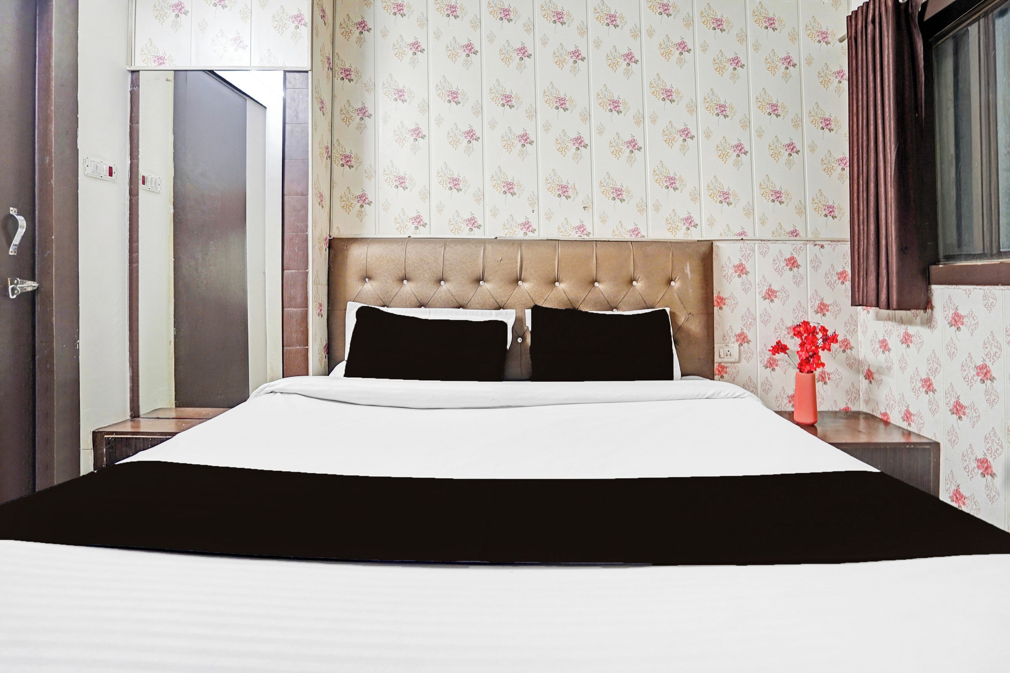 A hotel room with a bed, a nightstand, a vase with flowers, and a window with curtains.
