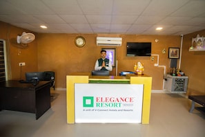 A reception desk at the Elegance Resort with a clock, air conditioning unit, and a TV on the wall.
