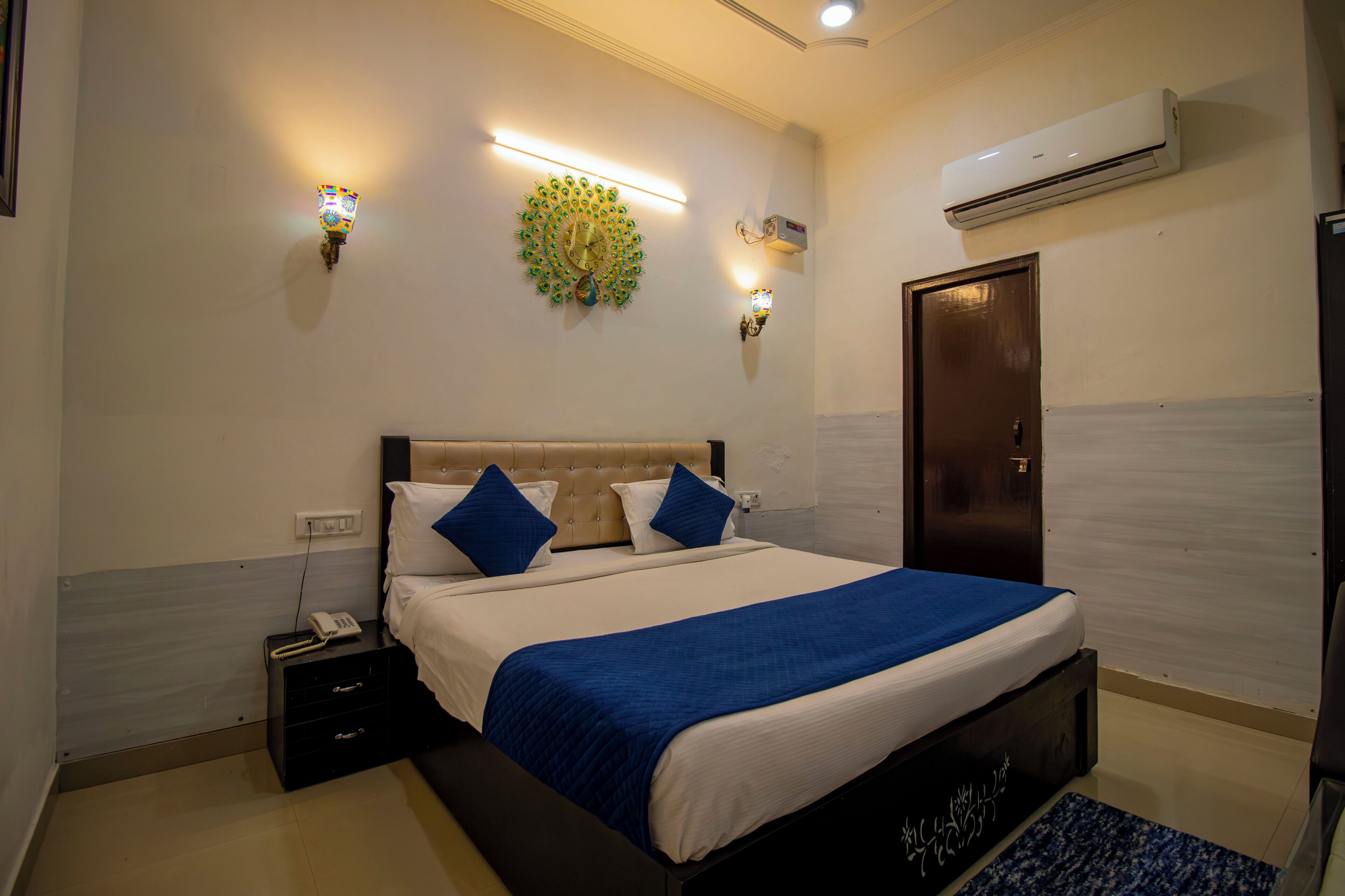 A hotel room with a bed, bedside tables, a telephone, wall-mounted lights, and an air conditioning unit.