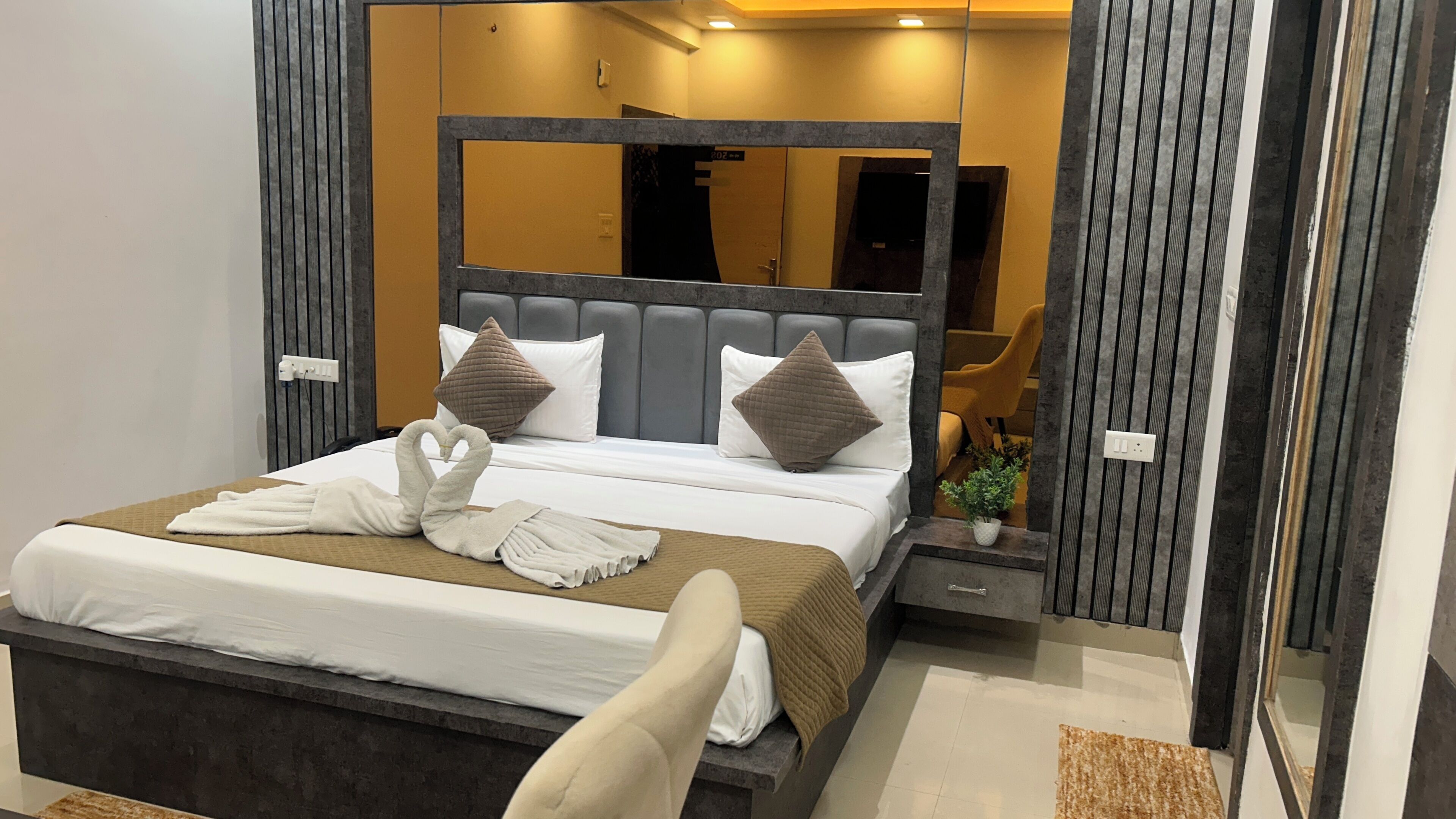 Una habitación de hotel moderna con una cama grande, cabecera con espejo, mesita de noche y una pequeña planta en maceta.