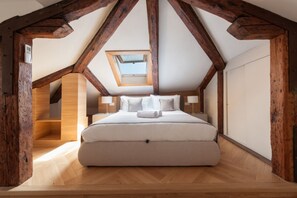 Ein Schlafzimmer mit schrägen Wänden, Holzbalken, einem Oberlicht, einem Bett mit Kissen und einem Nachttisch.