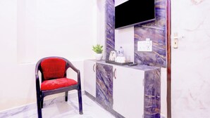 A room with a single red chair, a flat-screen TV mounted on a marble wall, a small cabinet with a plant, and a water dispenser. - Super OYO Collection O Subedarganj (Prayagraj)
