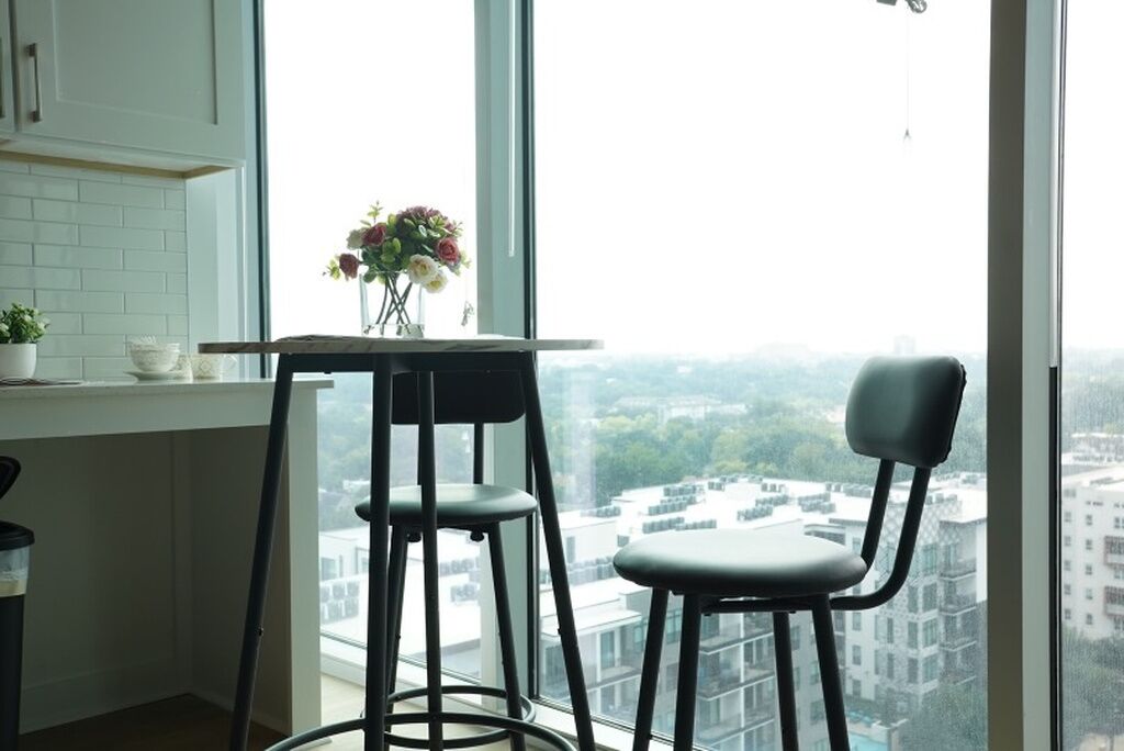 A kitchen with a window view of a cityscape, a table with a vase of flowers, and two bar stools.