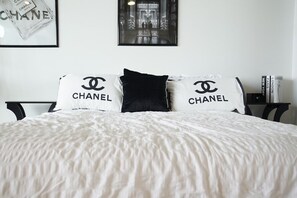 Ein Bett mit weißer Daunendecke und zwei Kissen mit 'CHANEL'-Branding, ein gerahmtes Bild über dem Bett und ein Nachttisch mit Büchern.