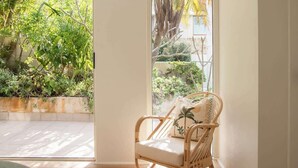 A room with a wicker chair, a bed with a green cover, and a view of a garden through a large window. - Aquamarine on Fairy Bower (Manly)