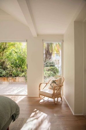 A room with a wicker chair, a bed with a green cover, and a view of a garden through a large window. - Aquamarine on Fairy Bower (Manly)