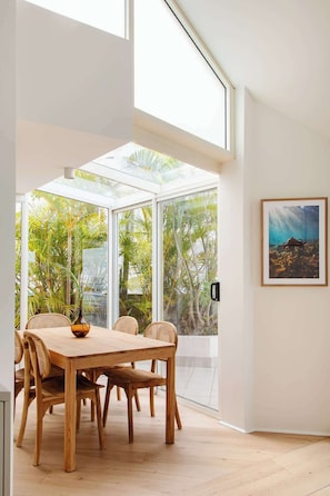 Un comedor con mesa y sillas de madera, ventanas amplias y un cuadro en la pared.