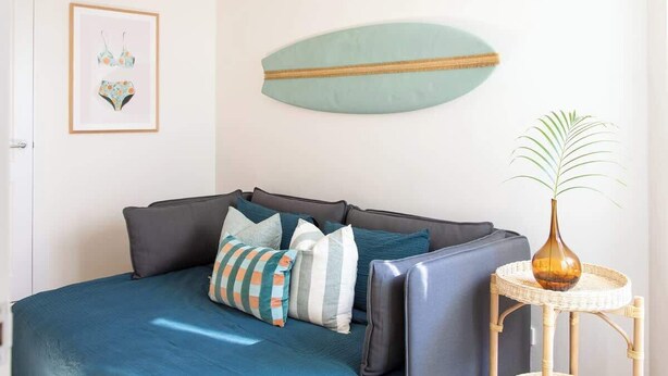 A room with a bed, pillows, a framed picture, a surfboard decoration, and a small table with a plant. - Aquamarine on Fairy Bower (Manly)