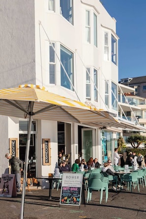 An outdoor seating area with tables and chairs, a signboard, and a building with large windows and a white facade. - Aquamarine on Fairy Bower (Manly)