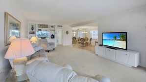 A living room with a white sofa, a television, and a dining area with chairs and a table. - Harborside Gardens at the Moorings (Naples)