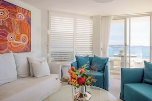 A modern living room with a white sofa, teal armchairs, a glass coffee table, and a colorful abstract painting on the wall. - Luxury Waterfront Living at Fairy Bower (Manly)