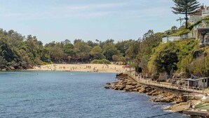 En kustvy med en strand, en klippig strandlinje, en träpromenad och en byggnad med en balkong som vetter mot stranden.