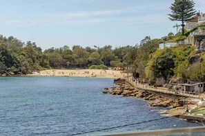 A coastal beach scene with a rocky shoreline, a wooden walkway, and a building with a balcony overlooking the beach. - Luxury Waterfront Living at Fairy Bower (Manly)