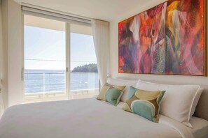 A hotel room with a large bed, a view of the sea through a sliding door, and a colorful abstract painting on the wall. - Luxury Waterfront Living at Fairy Bower (Manly)