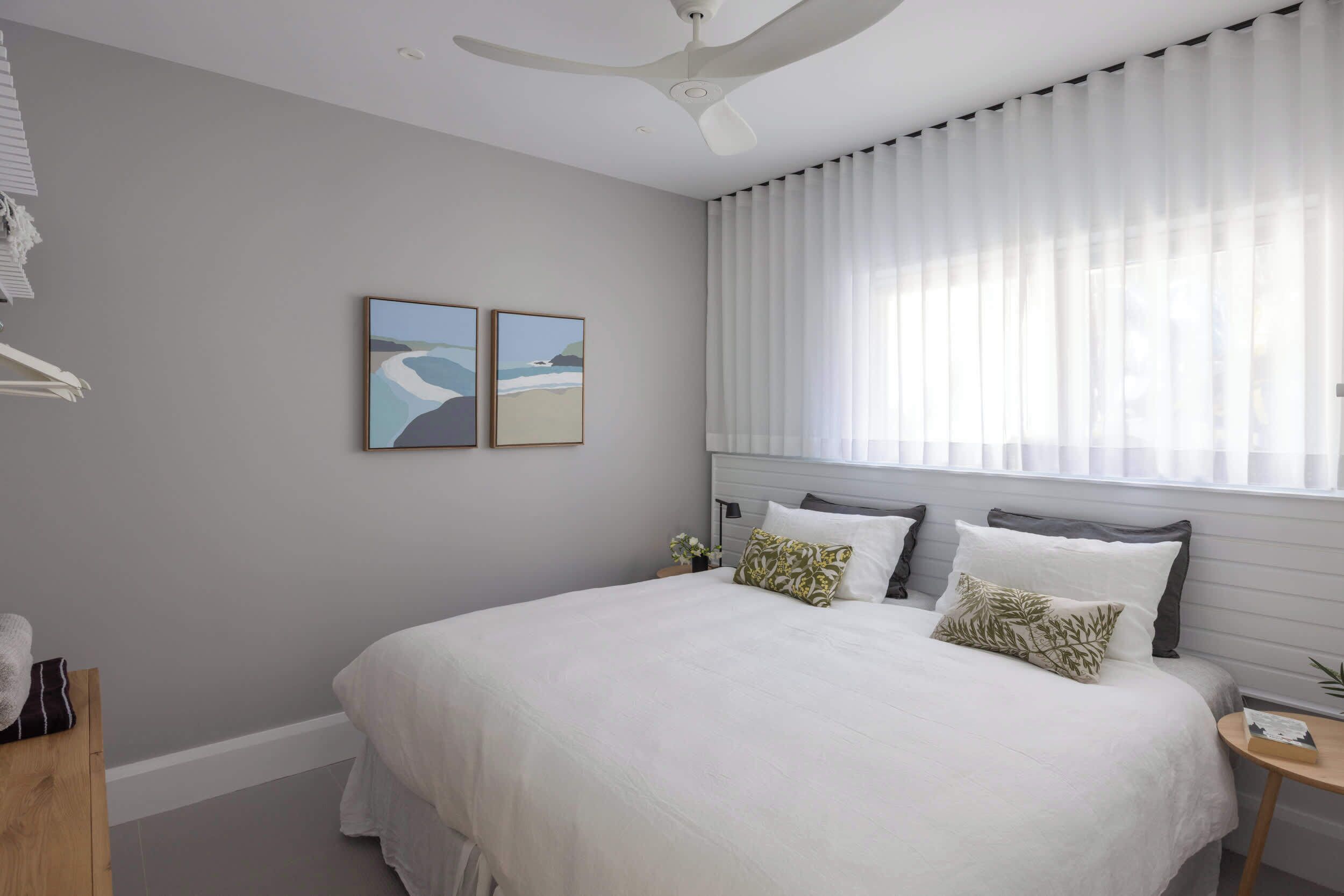 A bedroom with a large bed, a ceiling fan, a window with sheer curtains, and two framed pictures on the wall.