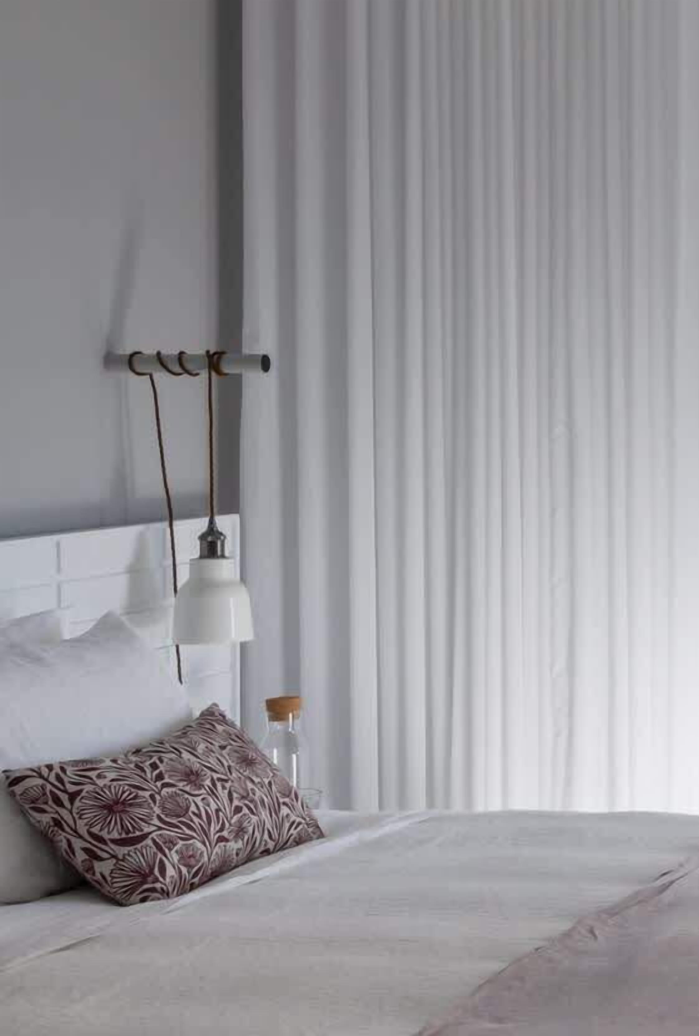 A neatly made bed with a patterned pillow, a wall-mounted lamp, and a window with vertical blinds.