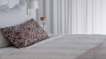 A neatly made bed with a patterned pillow, a wall-mounted lamp, and a window with vertical blinds.