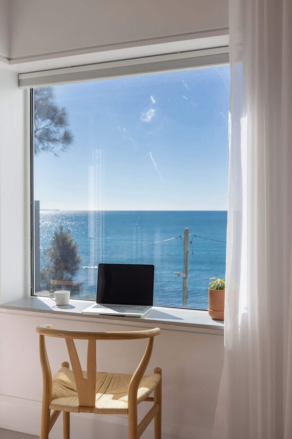 A laptop on a windowsill with a view of the sea and a chair in front.