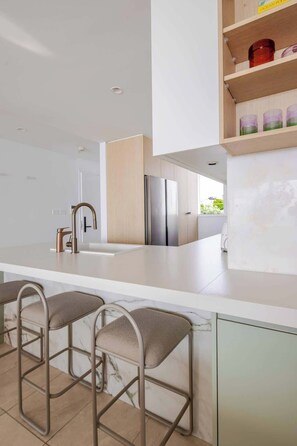 A modern kitchen with a central island, stainless steel appliances, and a bar area with stools. - Solaris - Oceanfront location with clifftop views (Vaucluse)