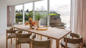 A modern dining room with a large table and chairs, a vase with flowers, and a view of a coastal landscape through large windows. - Solaris - Oceanfront location with clifftop views (Vaucluse)