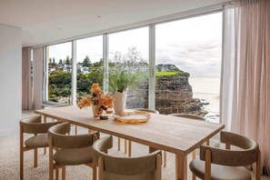 A modern dining room with a large table and chairs, a vase with flowers, and a view of a coastal landscape through large windows. - Solaris - Oceanfront location with clifftop views (Vaucluse)