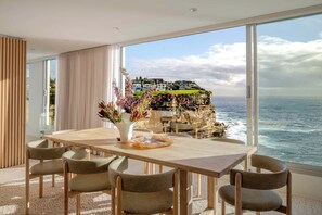 Ein Essbereich mit Tisch und Stühlen, einer Blumenvase und Blick auf das Meer sowie Häuser auf einer Klippe.