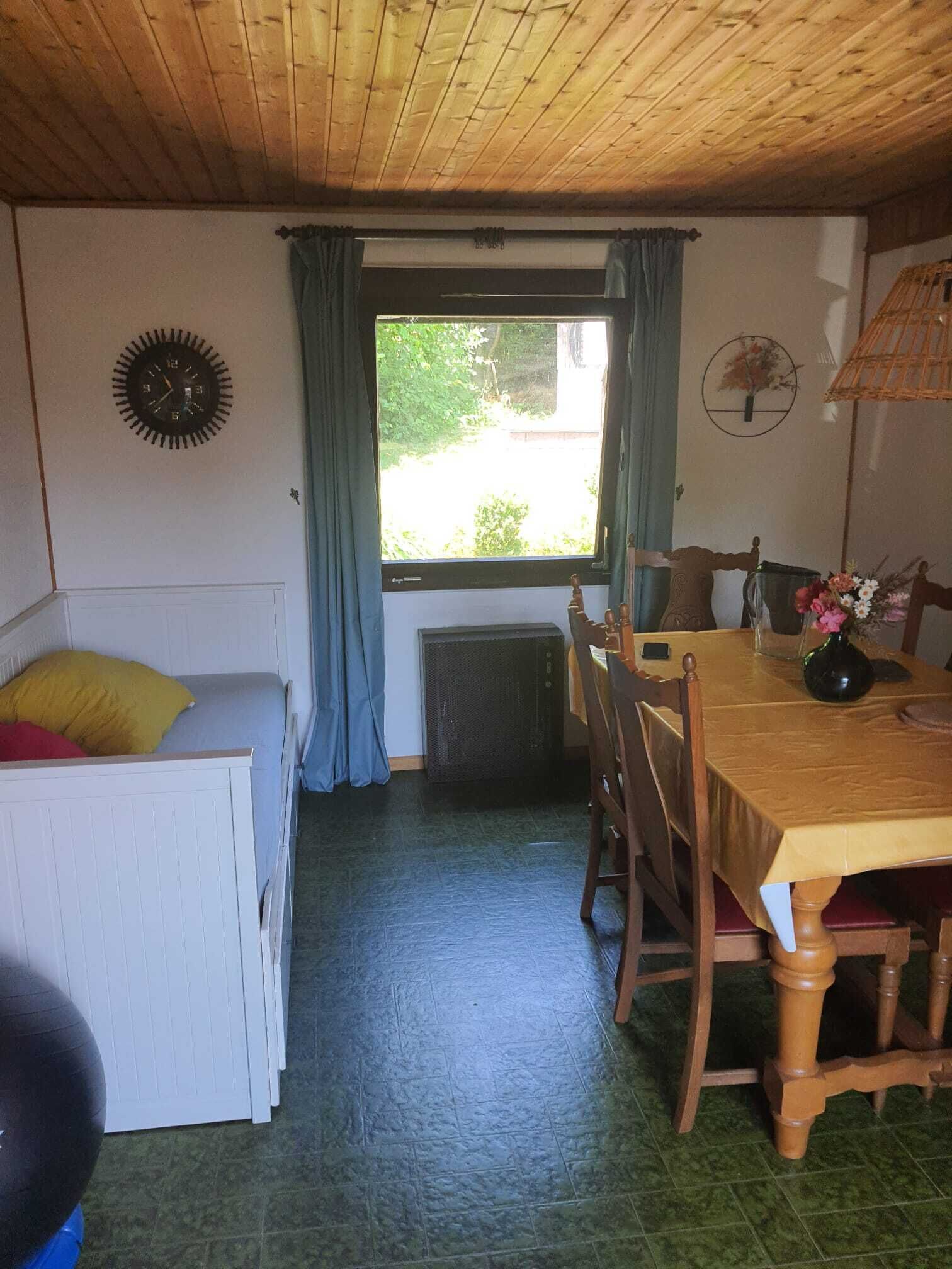 Huoneessa on sänky, ruokapöytä tuoleineen, ikkuna verhoineen ja patteri.