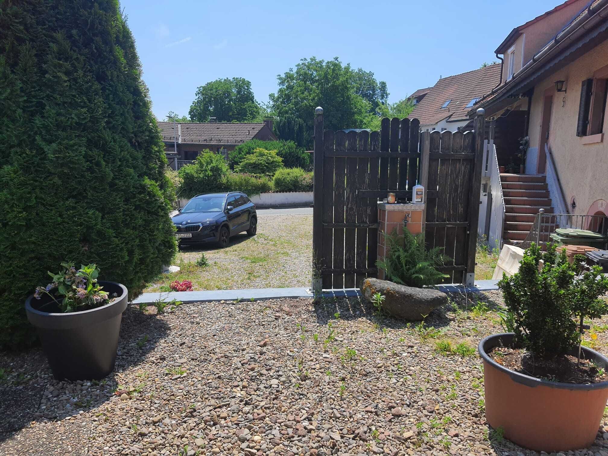 A gravel driveway leading to a house with a car parked in the driveway. There are potted plants and a wooden fence.