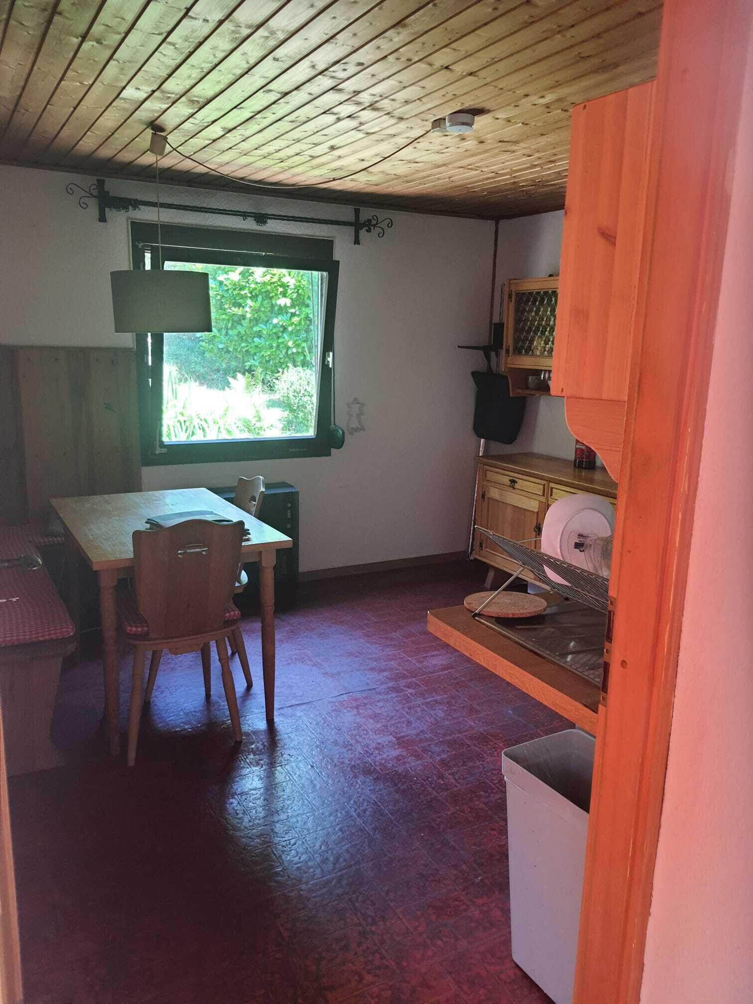 Une pièce avec un plafond en bois, une fenêtre donnant sur la verdure, une table avec des chaises et un petit meuble avec une étagère.