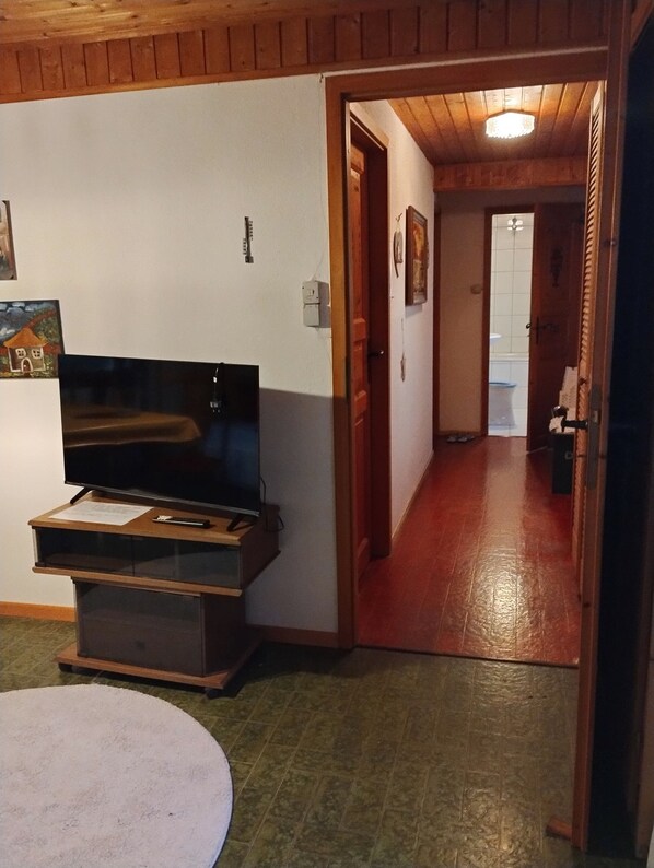 A room with a television on a wooden stand, a white rug, and a hallway leading to a bathroom.