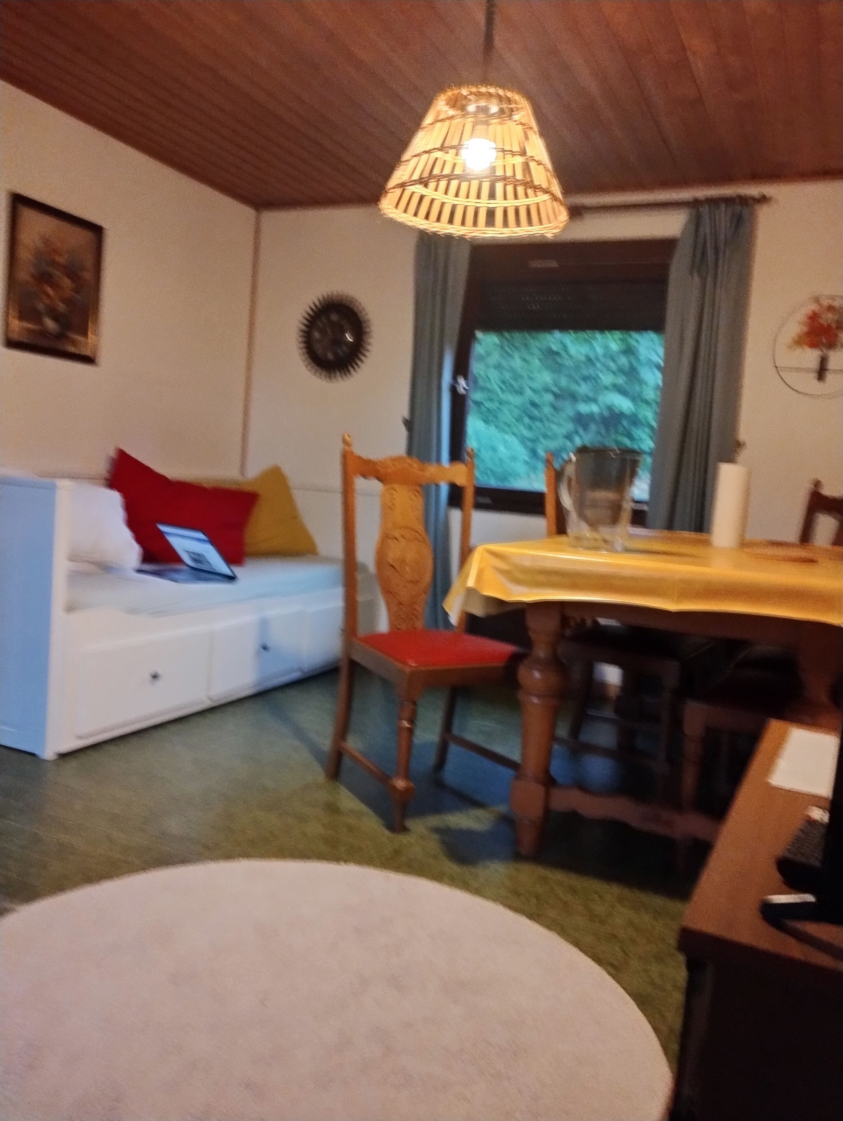 A room with a wooden ceiling, a white sofa with red and yellow cushions, a wooden chair, a dining table with a yellow tablecloth, and a window with curtains.
