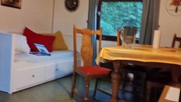 A room with a wooden ceiling, a white sofa with red and yellow cushions, a wooden chair, a dining table with a yellow tablecloth, and a window with curtains.