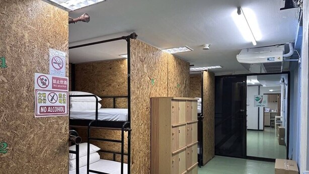 Habitación tipo dormitorio con literas, un armario de madera y un cesto de basura. Señales indican prohibido el consumo de alcohol y fumar.