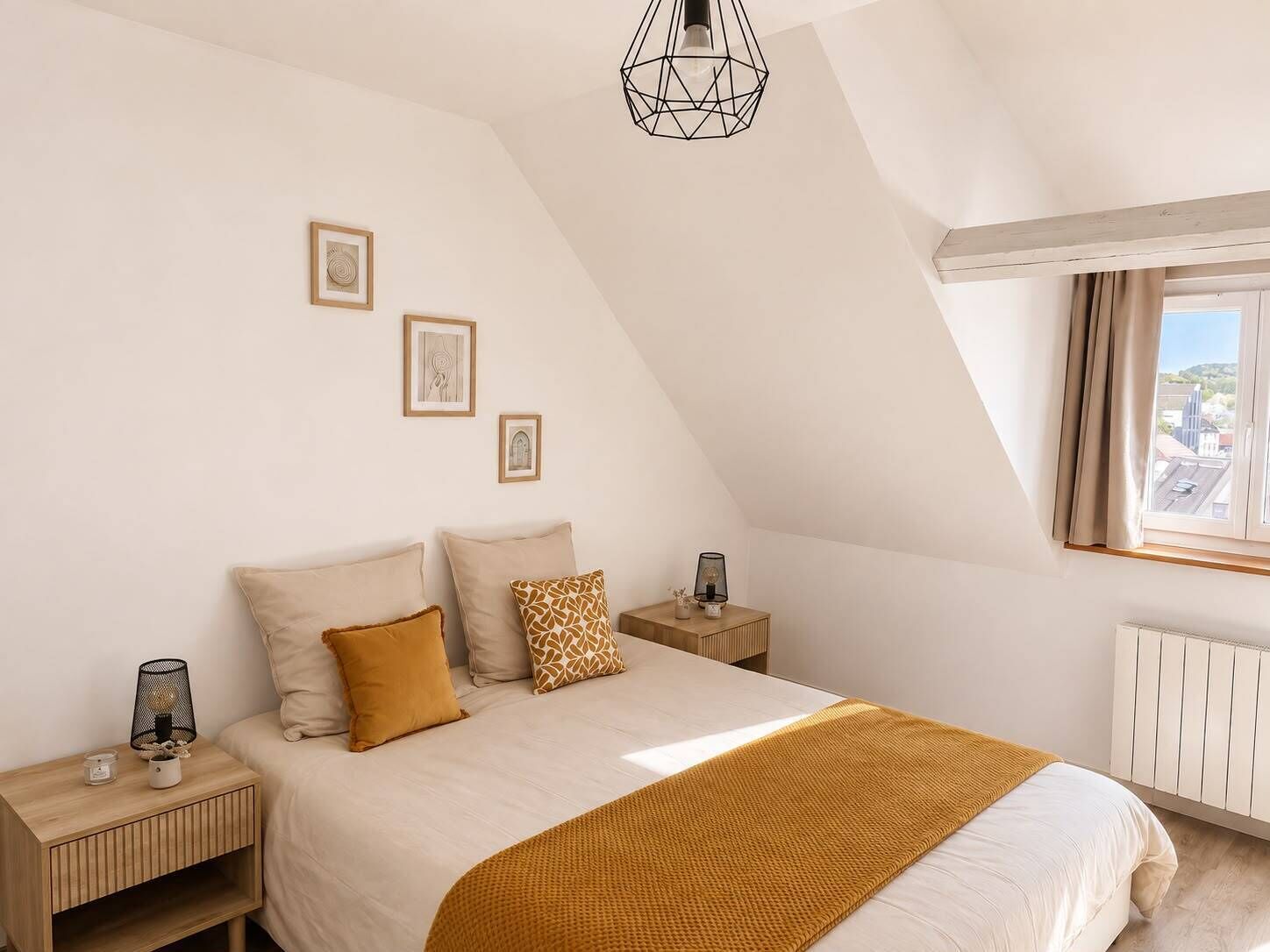 A bedroom with a sloped ceiling, a bed with a white comforter and a mustard-colored throw, a nightstand with a lamp, and a window with a view of buildings.