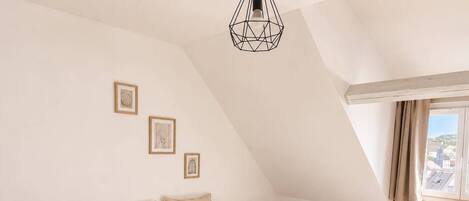Een slaapkamer met een schuine plafond, een bed met een witte dekbedovertrek en een mosterdkleurige plaid, een nachtkastje met een lamp, en een raam met uitzicht op gebouwen.