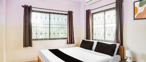 A bedroom with a bed, curtains, an air conditioning unit, and a bedside table with a lamp.