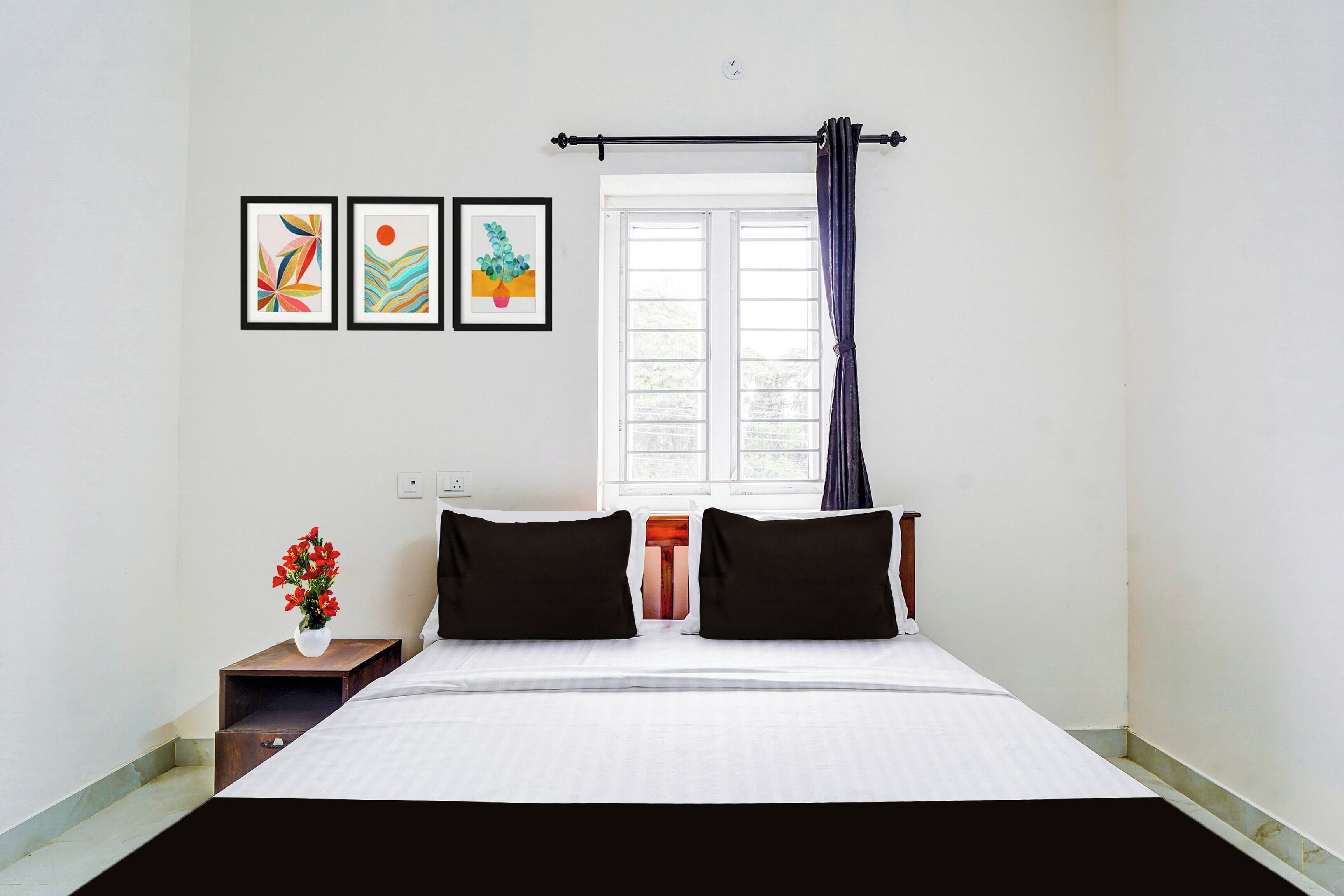A bedroom with a bed, two pillows, a nightstand with a vase of flowers, and three framed artworks on the wall.
