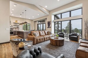A spacious living area with a large sofa, a coffee table, and a dining area with a kitchen in the background. - New Hilltop Escape, Sweeping Black Hills Views w/ Hot Tub, Large Driveway (Lead)