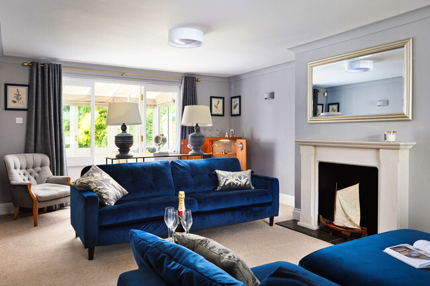 Een woonkamer met een blauwe bank, een grijze fauteuil, een open haard en een grote spiegel.
