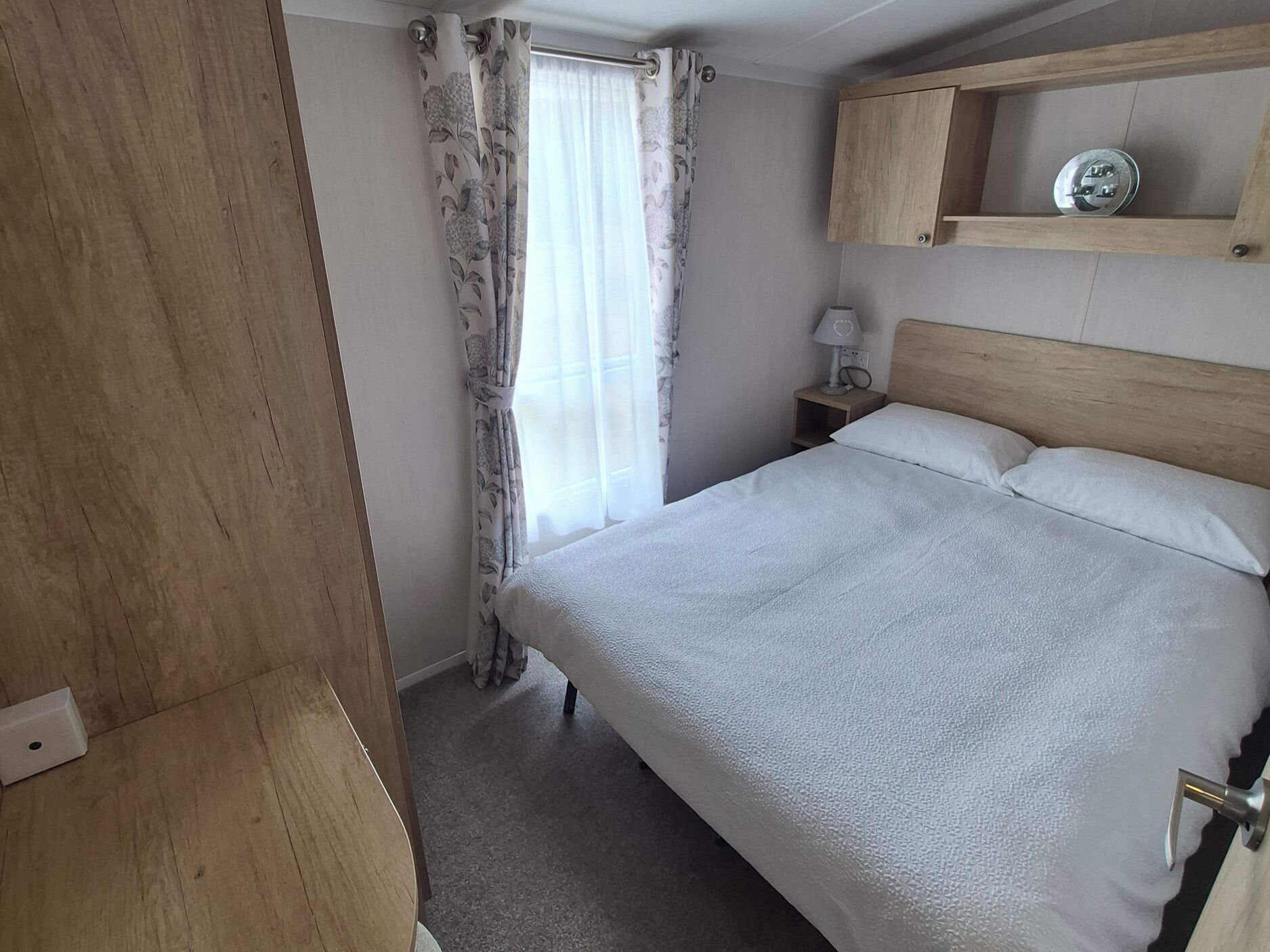 A bedroom with a bed, a nightstand, a lamp, a wooden shelf, and a window with curtains.
